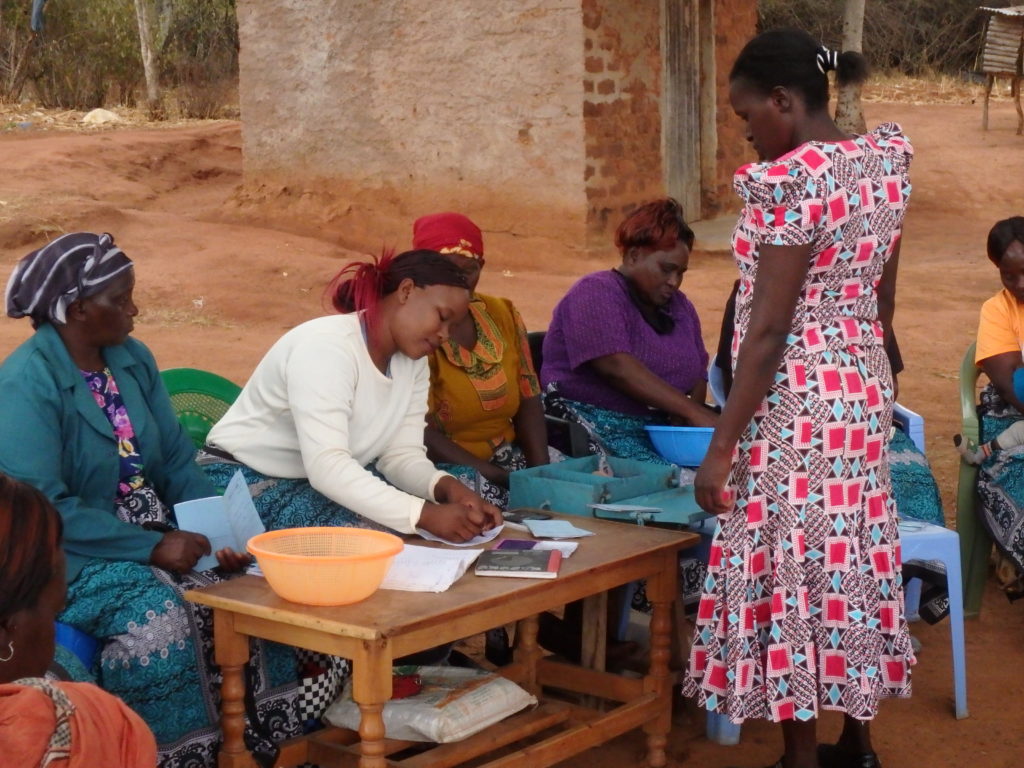 A group of women in a self-help group with one writing on a piece of paper while another woman waits for her to finish