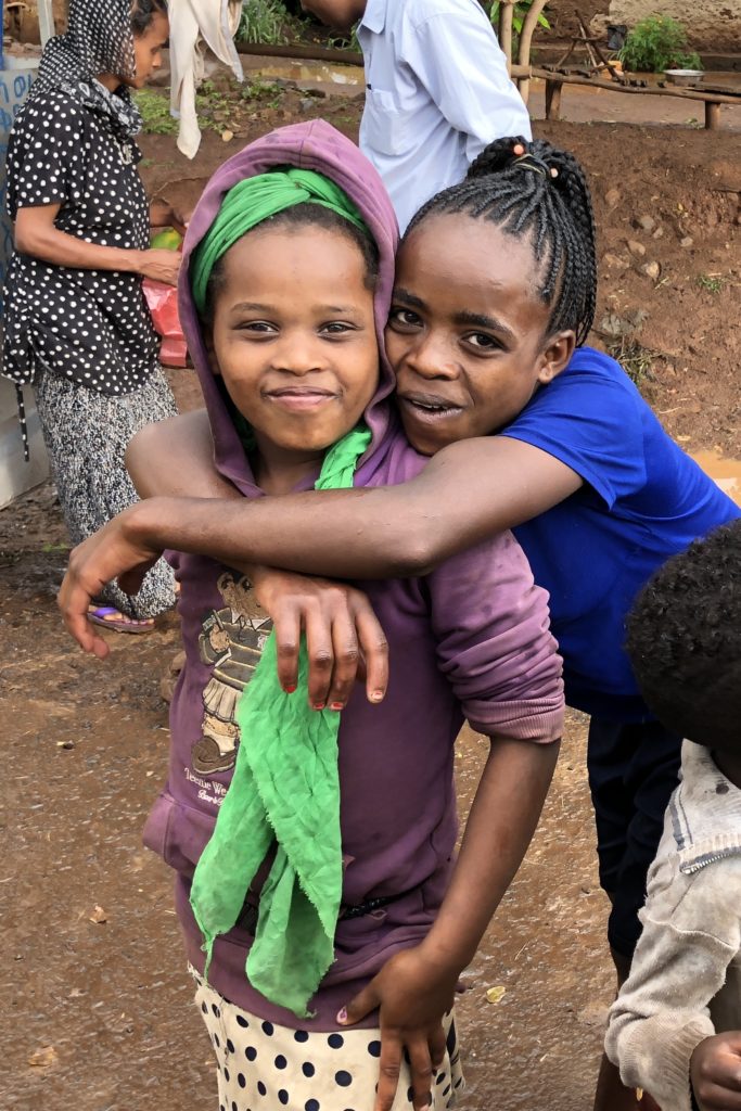 Two girls smiling at the camera. One girl has her arms around the other's shoulders, hugging.
