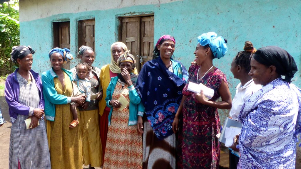 A group of women in a self-help group laughing together outside of a blue and white building. One woman is holding a young child.
