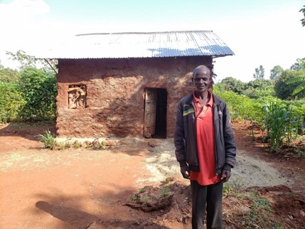 A man in front of a small one-bedroom mud house with a tin roof